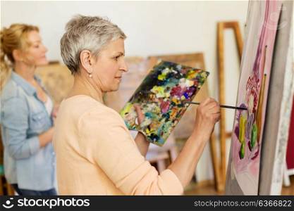 creativity, education and people concept - senior woman with brush and palette painting still life picture on easel at art school studio. senior woman painting at art school studio