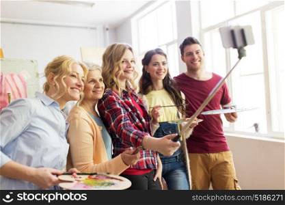creativity, education, technology and people concept - group of artists or students with brushes and palettes taking selfie by smartphone and monopod at art school studio. group of artists taking selfie at art school