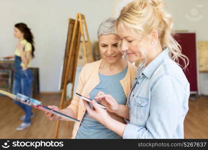 creativity, painting, technology and people concept - happy smiling women artists with smartphone, palette and easels at art school studio. artists with smartphone painting at art school