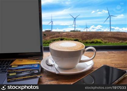 Credit card of laptop computer,smartphone and Hot coffee latte with latte art milk foam in cup mug on with wind turbines against and blue sky background,Online banking Concept