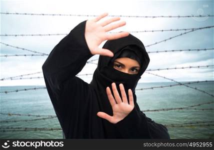 crime, imprisonment, violence, refugee and people concept - muslim woman in hijab making protective gesture over sea and barb wire background. muslim woman in hijab making protective gesture