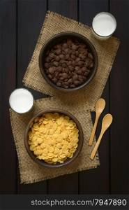 Crispy chocolate and simple corn flakes breakfast cereal in rustic bowls with glasses of milk and wooden spoons on the side, photographed overhead on dark wood with natural light