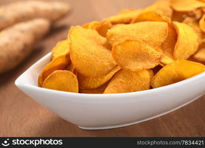 Crispy Peruvian sweet potato chips in white ceramic bowl with sweet potatoes in the back (Selective Focus, Focus one third into the sweet potato chips). Crispy Sweet Potato Chips