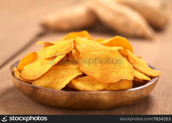 Crispy Peruvian sweet potato chips on wooden plate with sweet potatoes in the back (Selective Focus, Focus on the middle of the sweet potato chip in the front). Crispy Sweet Potato Chips