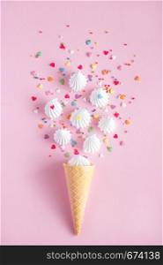 Crispy waffle ice creame cone with scattered white twisted meringues and with confectionary decorations on pink background