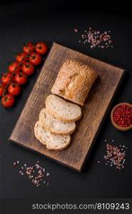 Crispy wheat flour baguette with sesame seeds on a wooden cutting board on a dark concrete background