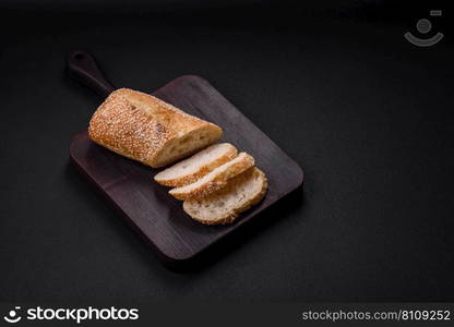 Crispy wheat flour baguette with sesame seeds on a wooden cutting board on a dark concrete background