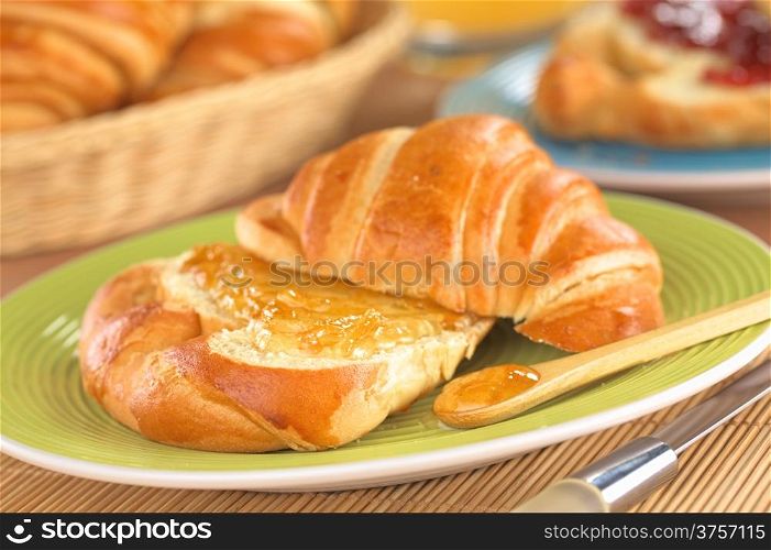 Croissant with butter and orange jam (Selective Focus, Focus on the front of the orange jam on the croissant and on the wooden spoon)