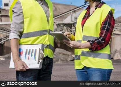 Crop anonymous female engineer showing information on tablet to male manager holding clipboard with diagrams while standing together near industrial structure and discussing project. Engineer and manager with tablet discussing project on industrial area