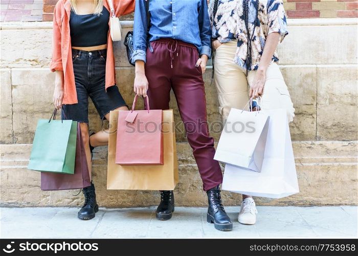 Crop anonymous stylish young female shopaholics in trendy outfits standing near aged stone building with shopping bags after successful purchases in city. Crop fashionable women with paper bags standing on street after shopping
