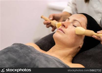 Crop beautician applying cosmetic cream on face of female customer with brushes during skin treatment in modern beauty center. Crop beautician massage with brushes on female face in beauty salon
