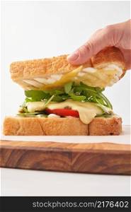 Crop man taking top toast of delicious sandwich with fresh green arugula and melted cheese over vegetables inside. Person removing toast off sandwich with vegetables
