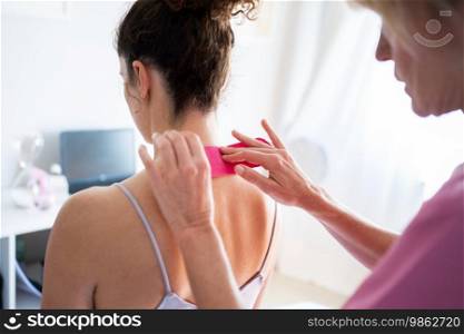 Crop professional osteopath applying kinesiology tape on neck of unrecognizable young female patient in inner wear during physiotherapy session in clinic. Woman applying soothing kinesiology tape on neck of client