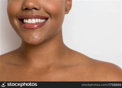 Crop young bare shouldered ethnic female model with perfect dark skin and lip gloss smiling brightly against white background. Happy ethnic woman with soft skin and toothy smile