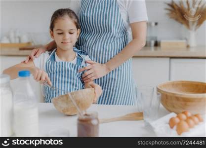 Cropped image of affectionate mother in apron embraces daughter who learns how to make pasty, holds whisk, mixes ingredients in bowl, poses at kitchen table with milk, melted chocolate, eggs