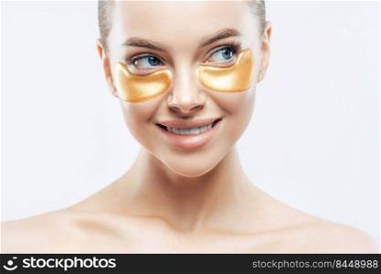 Cropped image of beautiful woman with naked shoulders, performs daily face care routine, applies golden patches under eyes to remove dark circles, isolated on white background. Beauty concept