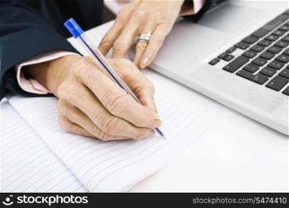 Cropped image of businesswoman with laptop writing in book on office desk