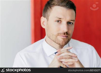 Cropped image of confident young male employee with bristle, keeps hands pressed together, dressed in elegant white shirt, contemplates about opening new firm, isoated over red and white wall