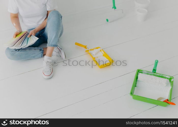 Cropped image of faceless woman chooses color from samples, going to refurbish walls in own room, wears sneakers, sits on white wall with trays and paint rollers. Repair and designing concept