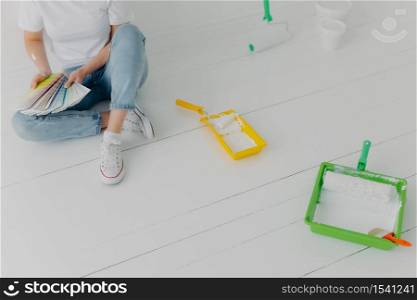 Cropped image of faceless woman chooses color from samples, going to refurbish walls in own room, wears sneakers, sits on white wall with trays and paint rollers. Repair and designing concept