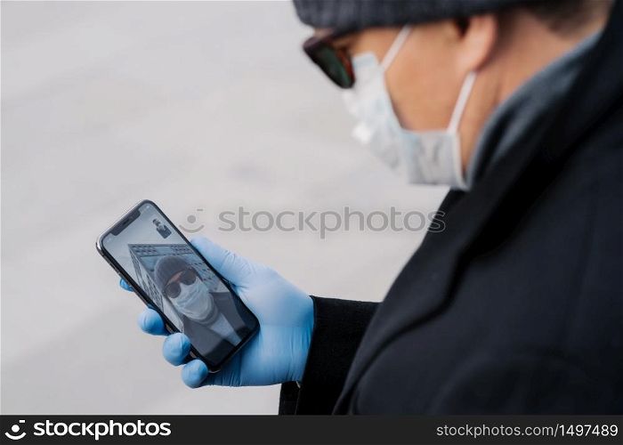 Cropped image of man has video call with best friend, keeps social distancing, being on self isolation during spreading infectious virus, wears protective mask and gloves, holds mobile phone in hand