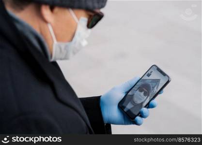 Cropped image of man has video call with best friend, keeps social distancing, being on self isolation during spreading infectious virus, wears protective mask and gloves, holds mobile phone in hand