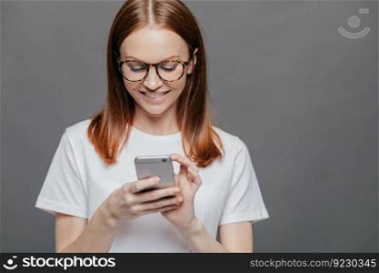Cropped image of pretty woman with cheerful expression, smiles broadly, holds mobile phone, happy to recieve message from boyfriend, stands against grey background with copy space on left side