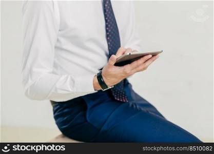 Cropped image of successful prosperous businessman in white shirt, black trousers, tie, holds modern tablet, checks notification, connected to wireless internet, isolated over white background