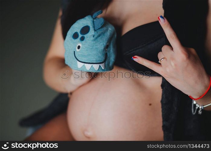 Cropped image pregnant woman holding hat for a baby in her hand and do rock symbol . Pregnant hugging tummy at home. Motherhood concept. Baby Shower. Cropped image pregnant woman holding hat for a baby in her hand and do rock symbol. Motherhood concept. Baby Shower
