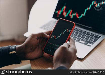 Cropped photo of male hands holding smartphone in front of computer screen analyzing financial stock market trading data charts, following investment growth graphs on phone and laptop monitors. Male hands holding smartphone next to computer screen analyzing financial stock market charts