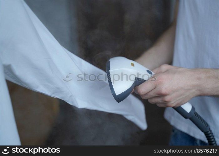 Cropped photo of man using steamer neatly ironing white shirt while holding long sleeve. Businessman tidies up formal outfit before important company meeting in office. Cropped photo of man using steamer neatly ironing white shirt while holding long sleeve