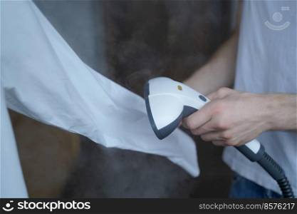 Cropped photo of man using steamer neatly ironing white shirt while holding long sleeve. Businessman tidies up formal outfit before important company meeting in office. Cropped photo of man using steamer neatly ironing white shirt while holding long sleeve