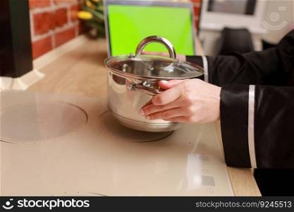Cropped photo of woman&rsquo;s hands puts metal cooking pan on induction stove at stylish home kitchen, laptop with green screen chroma key on table. Culinary blog, vlog, recipe for cooking.. Cropped photo of woman&rsquo;s hands puts metal cooking pan on induction stove at stylish home kitchen, laptop with green screen chroma key on table. Culinary blog, vlog, recipe for cooking