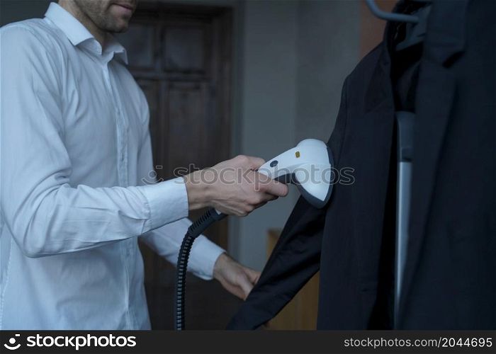 Cropped photo of young businessman preparing himself for important meeting with partner, cleaning his suit with portable travel steamer, experienced office worker steaming formal clothes in morning. Businessman preparing clothes for meeting with partner, steaming suit with portable steamer