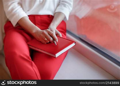 cropped photo of young European businesswoman with notebook is sitting by the window. Concept of planning. girl in white shirt and red pants. cropped photo of young European businesswoman with notebook is sitting by the window. Concept of planning. girl in white shirt and red pants.