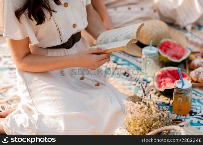 cropped photo of young woman in dress reading book while enjoying picnic in summer park. Sitting on blanket. Croissants, watermelon and cold summer drink.. cropped photo of young woman in dress reading book while enjoying picnic in summer park. Sitting on blanket. Croissants, watermelon and cold summer drink