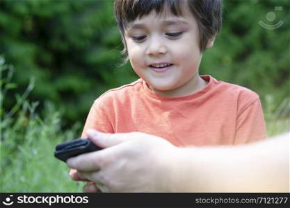 Cropped shot Kid boy siting in the park with parent playing game on smart phone, Child looking at mobile phone with blurry green nature background. happy family concept