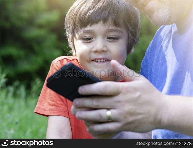 Cropped shot Kid boy siting in the park with parent playing game on smart phone, Child looking at mobile phone with blurry green nature background. happy family concept