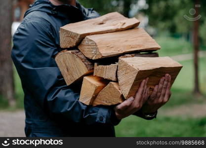 Cropped shot of faceless man in black jacket carries pile of firewood poses against blurred forest background.