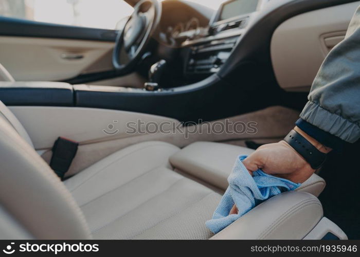 Cropped shot of man cleaning car interior, wiping fabric passenger seat with microfiber cloth, removing dirt and dust in modern vehicle salon with rag. Car wash concept. Man cleaning car interior with microfiber cloth at carwash service