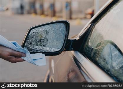 Cropped shot of man cleaning car outdoors with rag, wiping water droplets with microfiber cloth on side rearview mirror of auto during vehicle washing at self-service station. Car wash concept. Male hand cleaning car outdoors with rag, wiping water with microfiber cloth on side rearview mirror