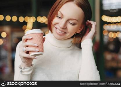 Cropped shot of pleasant looking woman poses in outdoor cafe, holds paper cup of coffee, dressed in white outfit, smiles happily, has talk with colleague or friend during break. Female with beverage