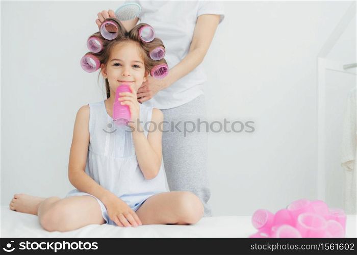Cropped shot of pleased little child with curlers on head and her mother stands behind combs hair and winds curlers, have appealing appearnce. People, loving family and relationship concept.