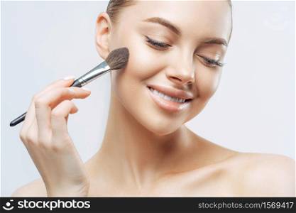 Cropped shot of positive European woman with healthy soft skin, uses cosmetic powder brush, applies foundation on face, has pure clean complexion, natural makeup, stands indoor. High resolution