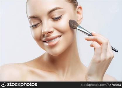 Cropped shot of positive European woman with healthy soft skin, uses cosmetic powder brush, applies foundation on face, has pure clean complexion, natural makeup, stands indoor. High resolution