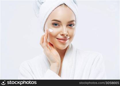 Cropped shot of pretty young woman applies cream for rejuvenation, healthy soft skin, uses cosmetic product, demonstrates nice effect of body lotion, wears comfortable soft white bath robe, towel