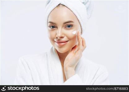 Cropped shot of pretty young woman applies cream for rejuvenation, healthy soft skin, uses cosmetic product, demonstrates nice effect of body lotion, wears comfortable soft white bath robe, towel