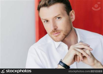 Cropped shot of serious young male CEO or company owner, has success, climbs career ladder, looks with strict expression at camera, keeps hands together, poses against red and white background