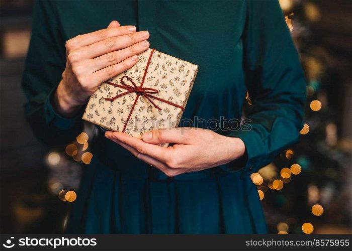 Cropped shot of unrecognizable woman in festive dress being glad to recieve christmas present from husband stand against New Year decorated fir tree. Congratulation and celebration concept