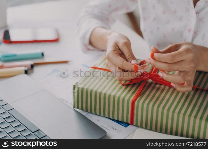 Cropped shot of woman online store business owner packing package gift box for customer during Christmas winter holidays, female tying red bow on xmas present, preparing parcel on table. Female hands packing package gift box for customer during Christmas winter holidays